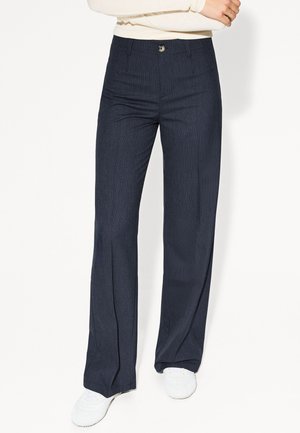 Persoon die donkerblauwe wijde broek met krijtstrepen en witte sneakers draagt, met gekruiste armen over een witte top met lange mouwen.