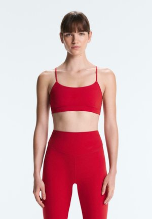 Ženska s ravnimi rjavimi lasmi, oblečena v oprijet rdeč športni modrček in hlače z visokim pasom, stoji pred svetlo sivo ozadje.