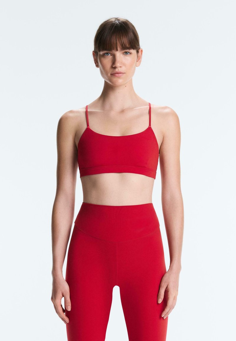 Kobieta o prostych brązowych włosach, ubrana w dopasowany czerwony sportowy stanik i legginsy z wysokim stanem, stojąca na tle jasnoszarego tła.