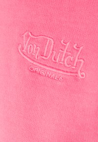 Roza bombažna tkanina z vezenim logotipom "Von Dutch Originals" v teksturiranem slogu. Poudarjen bližnji posnetek prikazuje gladko tekstilno površino in črke.