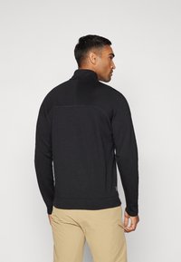 Mustakirja, pika varrukaga pullover kõrge kraega ja peene tekstuuriga. Omab õmblust ülemise seljaosas ja soonikkoes mansetid, millele on kombineeritud beežid püksid.