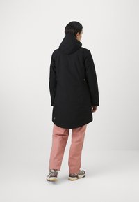 Zwarte waterdichte jas met capuchon, lange stijl, zijzakken en blauwe accenten. Draagt over lichtroze broek en lichte schoenen.