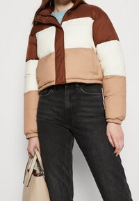 Puchowa kurtka z poziomymi blokami kolorów w brązie, beżu i bieli. Materiał jest błyszczący, z cropped designem i zamkiem z przodu.