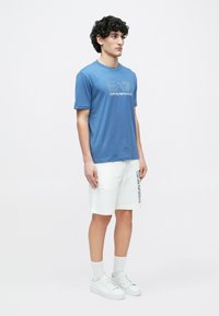 Mees, kes seisab seljas sinine EA7 Emporio Armani t-särk, valged lühikesed püksid logoga, valged sokid ja valged tossud, ühevärvilisel taustal.