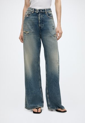 Jeans blu scoloriti a gamba larga con dettagli consumati indossati con sandali neri e top bianco senza maniche, mostrati dalla vita ai piedi.