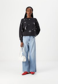 Zwarte trui met witte ster- en maanpatronen, gecombineerd met lichtblauwe wijde jeans, rode instappers en een witte handtas.
