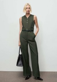 Mono verde oliva con escote en V, cierre de botones y bolsillos laterales; estilizado con un cinturón negro y un bolso negro; textura suave.