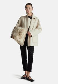 Witte wollen jas met een brede kraag, zijzakken en oversized pasvorm. Gecombineerd met zwarte broeken en zwarte platte schoenen. Houdt een fluffy beige tas vast.