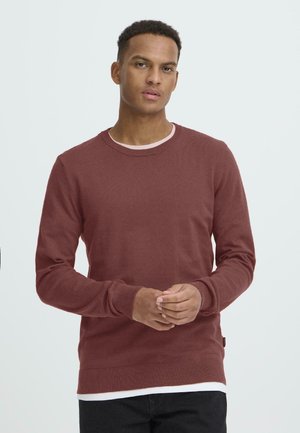 Bordeauxroter Langarmpullover aus weichem Strickmaterial, mit Rundhalsausschnitt, gerippten Bündchen und einem leicht lockeren Schnitt. Über einem weißen T-Shirt getragen.