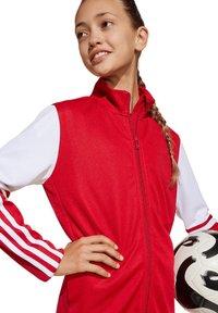 Rote Sportjacke mit weißen Ärmeln, Reißverschluss vorne und drei weißen Streifen an den Ärmeln. Hält einen Schwarz-Weiß-Fußball.