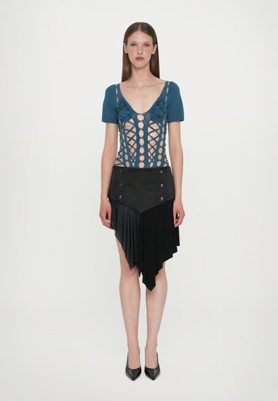 Body bleu découpé avec des ouvertures circulaires, associé à une jupe plissée noire asymétrique avec des boutons et une texture lisse. Talons noirs.