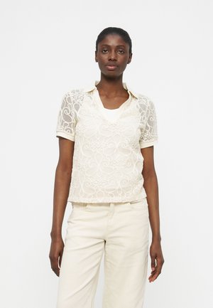 Γυναίκα που φοράει πουκάμισο πόλο με κοντά μανίκια και κρεμ δαντέλα πάνω από λευκό τοπ, με κρεμ παντελόνι υψηλής μέσης, στέκεται μπροστά σε λευκό φόντο.