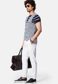 Mężczyzna w szarym w kratę kamizelce nałożonej na granatową w paski koszulę, białych spodniach i ciemnych sneakersach, trzymając brązową skórzaną torbę.