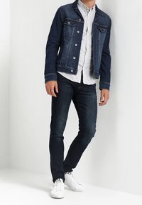 Ciemna kurtka jeansowa o zorganizowanym kroju, z zapięciem na guziki z przodu i dwoma kieszeniami na piersi, zestawiona z ciemnymi dżinsami i białymi trampkami.