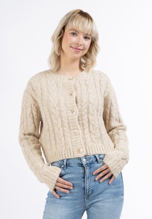 Jonge vrouw met blond haar die een beige kabelgebreide, geknoopte cardigan en lichtblauwe spijkerbroek draagt tegen een effen witte achtergrond.