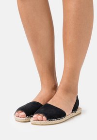 Svart mocka espadrille-lägenheter med öppen tå, en vävd jutesula och sömnadsdetaljer. Slät textur, minimalistisk design och låg profil.