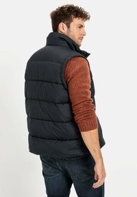 Navy gepolsterte Weste mit hohem Kragen, die über gesteppten Bereiche verfügt. Gestylt mit einem strukturierten orangefarbenen Strickpullover und blauen Jeans.