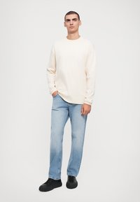 Long-sleeve cream t-shirt, relaxed fit, paired with light blue straight-leg jeans. Black lace-up shoes complete the outfit. White background.