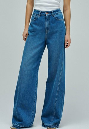 Mujer vestida con vaqueros azules de talle alto y pierna ancha y una camiseta blanca sin mangas, de pie contra un fondo gris claro liso.