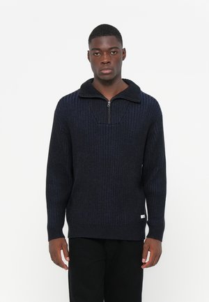 Suéter de punto acanalado azul oscuro con un alto cuello y media cremallera, que presenta una tela texturizada y una etiqueta en el dobladillo.