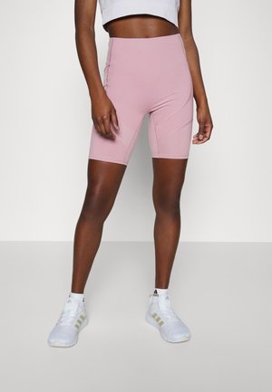 Kobieta w różowych rowerowych szortach z wysokim stanem i białych butach sportowych Adidas na jednolitym szaro-białym tle.