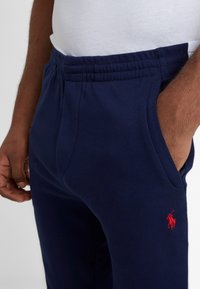 Mörkblå bomulls-joggers med elastisk midja, sidofickor och röd broderad logotyp. Mjuk textur, avslappnad passform och enkel design.