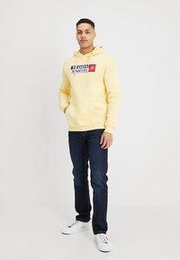 Gele hoodie met groot "Teddy Smith" logo op de voorkant, gecombineerd met donkere spijkerbroeken en witte sneakers. Zachte stof, met kangoeroezak detail.
