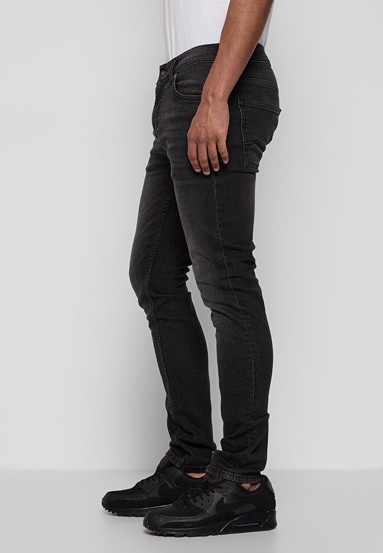 Cienkie czarne dżinsy wykonane z denimu, z klasycznym pięciopocketowym wzorem, subtelnym przeszyciem i gładką fakturą. Noszone z czarnymi sneakersami.
