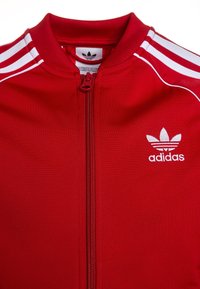Červená bunda Adidas, se zipem vpředu, vysokým límcem, bílými pruhy na ramenou a vyšitým logem na levé části hrudi. Hladká textilie.