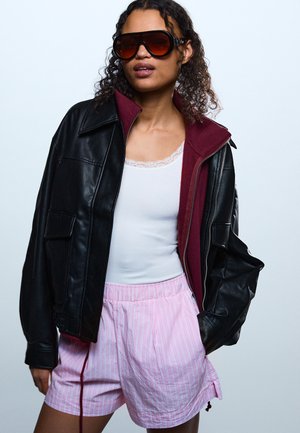 Γυναίκα που φορεί υπερμεγέθη μαύρο δερμάτινο μπουφάν, μπορντό φούτερ με φερμουάρ, λευκό αμάνικο τοπ, ροζ ριγέ σορτς και μεγάλα σκούρα γυαλιά ηλίου.