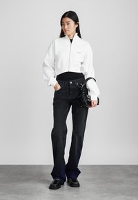 Witte cropped zip-up sweatshirt over een zwarte top, gecombineerd met zwarte flared jeans. Accessoires zijn een kleine zwarte tas en zwarte schoenen.