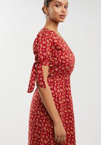 Rdeča cvetna obleka s kratkimi nabranimi rokavi, zavezanimi v pentlje. Ima prilegajoč bodice in tekočo krilo z majhnimi rumenimi in belimi cvetličnimi vzorci.