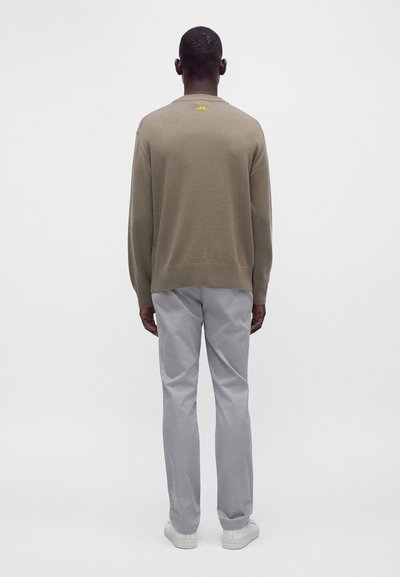 Homme debout, de dos, portant un pull taupe avec un petit logo jaune sur le haut du dos, un pantalon gris clair et des baskets blanches.