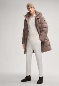 Cappotto puffer marrone con colletto alto, camicia crema, pantaloni bianchi e stivali in suede scuri. Berretto in maglia beige con dettaglio del logo.