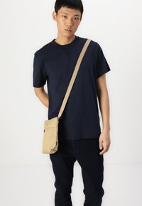Tricou bleu cu mânecă scurtă, geantă bej cu umăr și buzunar frontal, blugi închiși la culoare. Design simplu, material natural, estetică minimală.