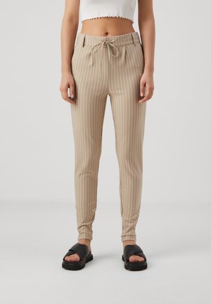 Pantalon beige à rayures avec une taille à cordon, coupe ajustée et poignets élastiques. Porté avec des sandales à glissière noires.