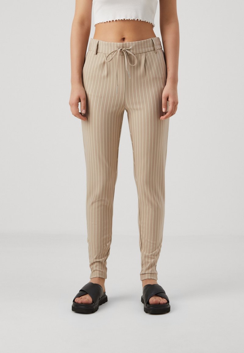 Pantalon beige à rayures avec une taille à cordon, coupe ajustée et poignets élastiques. Porté avec des sandales à glissière noires.