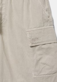 Tissu beige à texture quadrillée, avec une poche à rabat brodée d'un petit logo de marque en fil assorti.