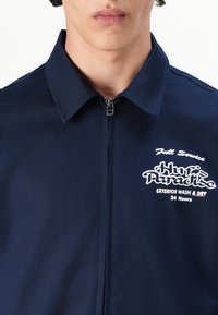 Navy blue zip-up jacket with a collar, featuring white embroidered text "Huf's Paradise" and "Exterior Wash & Dry 24 Hours." Textured fabric.