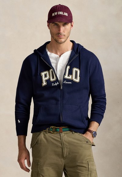 Tummansininen zip-up huppari, jossa on brodeerattu "POLO" logo, valkoinen t-paita alla, oliivinvihreät cargo-housut ja viininpunainen lippislakki, jossa on "NEW ENGLAND."