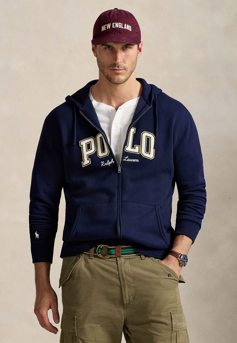 Mörkblå zip-up hoodie med broderad "POLO"-logga, vit t-shirt under, olivgröna cargobyxor och en vinröd keps med "NEW ENGLAND."