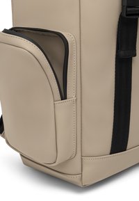 Beige hátizsák, textúrált külsővel, oldalsó cipzáras zsebbekkel és fekete díszítésekkel, beleértve a pántokat és a varrásokat. Simaság és minimalista dizájn jellemzi.