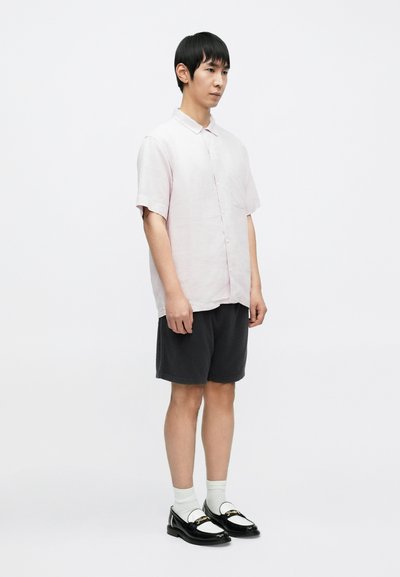 Κοντομάνικο, ανοιχτό ροζ λιναρό πουκάμισο, γκρι σορτς, λευκές κάλτσες, μαύρα loafers με λευκές λεπτομέρειες και χρυσά μεταλλικά στοιχεία, που στέκεται σε απλό φόντο.