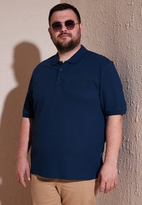 Jūros mėlynos spalvos trumpomis rankovėmis polo marškiniai su klasikiniu apykakle ir trimis sagomis, dėvimi kartu su chaki spalvos kelnėmis, prieš faktūruotą sienos fono.