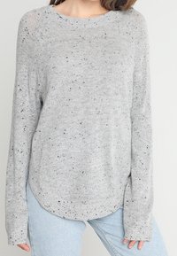 Grauer Strickpullover mit schwarzen Sprenkeln, runder Halslinie, langen Ärmeln und abgerundetem Saum, kombiniert mit hellblauen Jeans.