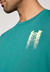 Τυρκουάζ κοντομάνικη μπλούζα κατασκευασμένη από απαλό ύφ ασμα. Διαθέτει κίτρινο λογότυπο Under Armour στο στήθος με εφέ διαβάθμισης. Λείος υφή.