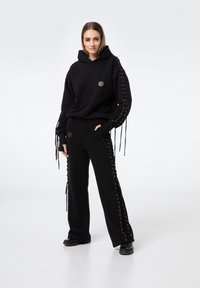 Czarna bluza z kapturem z detalami sznurowania na rękawach, w połączeniu z dopasowanymi czarnymi spodniami z koronkowymi akcentami po bokach. Oba elementy wykonane z miękkiego materiału.