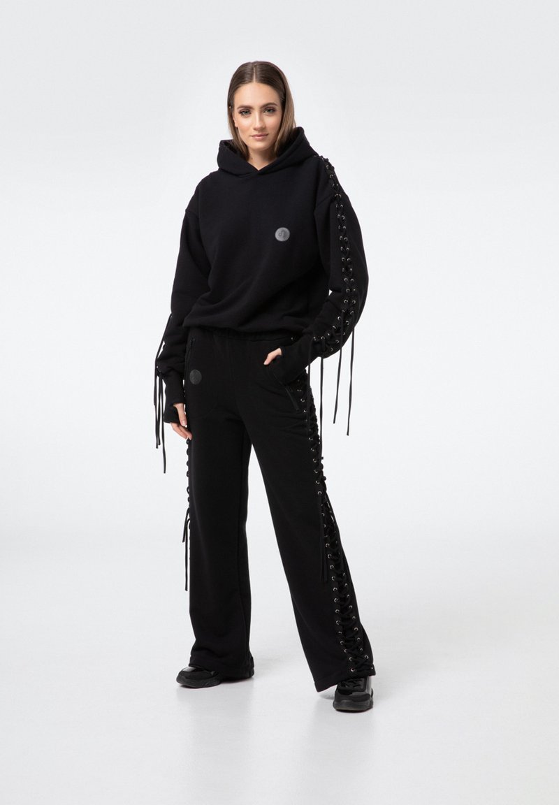 Czarna bluza z kapturem z detalami sznurowania na rękawach, w połączeniu z dopasowanymi czarnymi spodniami z koronkowymi akcentami po bokach. Oba elementy wykonane z miękkiego materiału.
