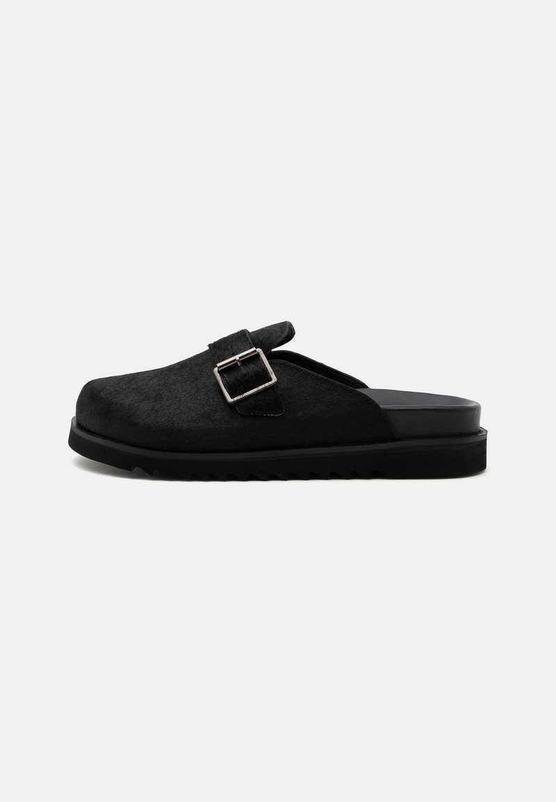Sabot noir à enfiler avec dessus texturé, sangle à boucle argentée et semelle épaisse rainurée, présenté en profil latéral sur fond blanc.
