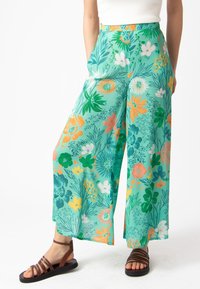 Brede bloemenbroek in groen, met een mix van oranje, witte en gele bloemen, gemaakt van lichte stof en voorzien van zijzakken.